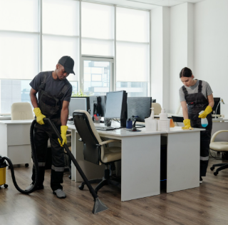 Two professional cleaners using equipment to clean an office space with computers and desks.