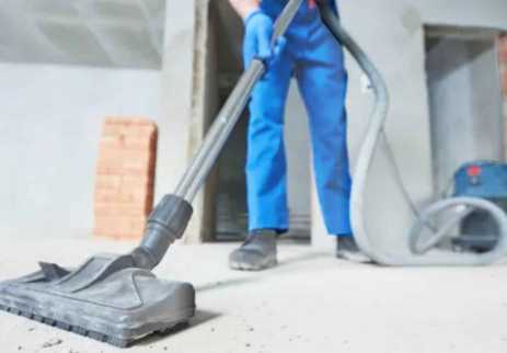 Person in blue overalls using a vacuum cleaner on a construction site floor.