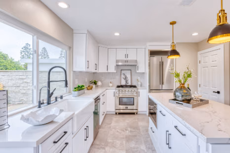 Modern kitchen with white cabinets, stainless steel appliances, and marble countertops featuring a large window and pendant lighting.