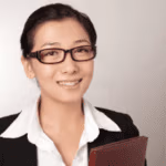 Smiling woman in a black blazer and glasses holding a folder, with a neutral background.